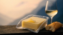Cheese in plastic packaging tray on a wooden table with white bread and a glass of wine, set against a scenic mountain view in soft evening light. Cheese in plastic packaging tray on a wooden table with white bread and a glass of wine, set against a scenic mountain view in soft evening light.